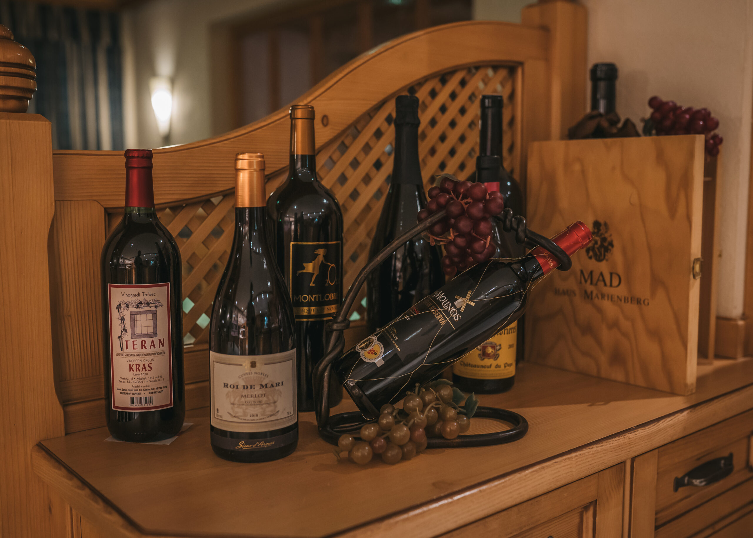 Eine sorgfältig ausgewählte Weinsammlung, darunter Flaschen von Teran und Roi de Mari Merlot, arrangiert auf einem Holztisch neben Weintrauben