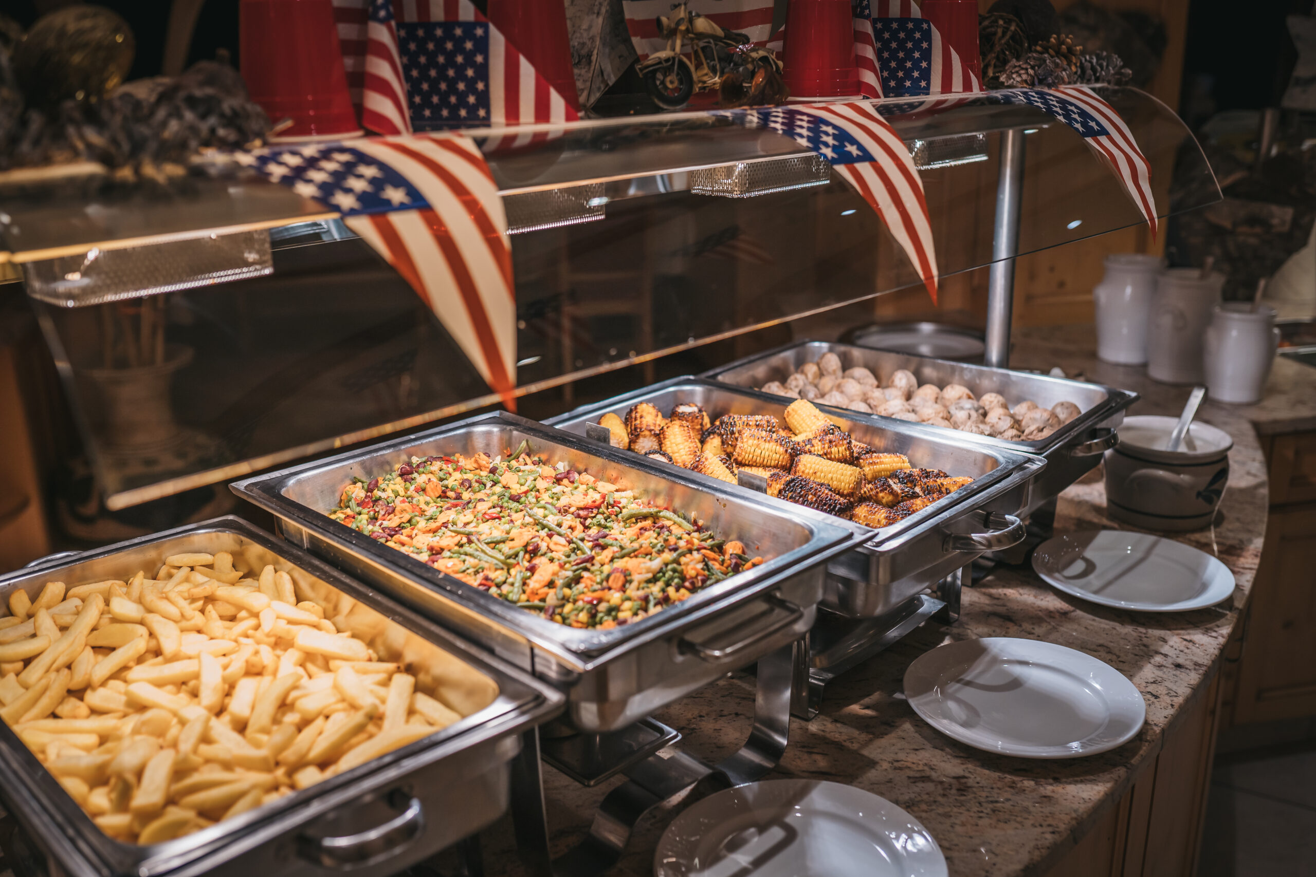 Ein Buffet mit Pommes, gegrilltem Mais und einer Vielzahl von Gemüsegerichten, dekoriert mit amerikanischen Flaggen im Hintergrund