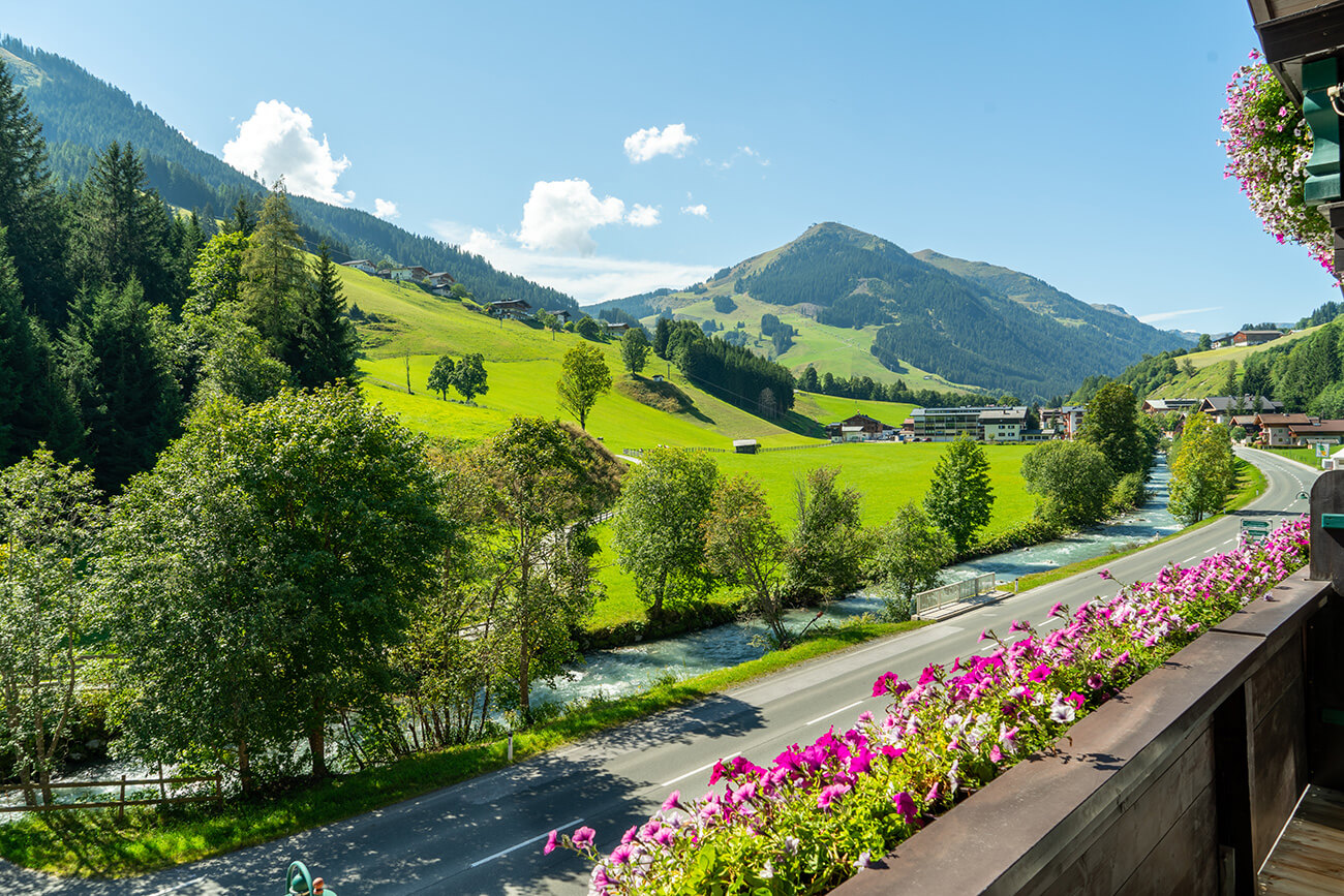 Idyllischer Blick von einem Hotelbalkon, geschmückt mit pinken Blumen, der eine Panoramaaussicht auf einen friedvollen Fluss, ein üppiges grünes Tal und sanfte Hügel