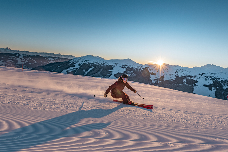 Skifahrer in roter Jacke gleitet bei Sonnenaufgang über eine perfekt präparierte Piste mit beeindruckendem Bergpanorama in Saalbach Hinterglemm.