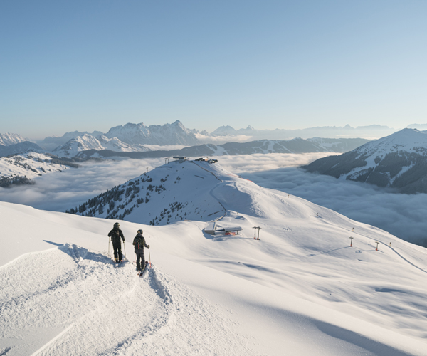 Zwei Wanderer in Winterausrüstung besteigen einen verschneiten Bergrücken mit Blick auf ein tief liegendes Nebelmeer und Berggipfel im Hintergrund.